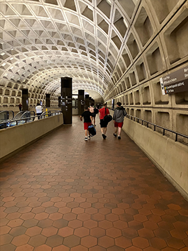 A picture of a pathway in a metro station. To the left and right there are two side walls with railing on top. The wall on the left ends in the background and has a wayfinding structure at the end. In the center of the picture in the background there is a family of four with luggage.